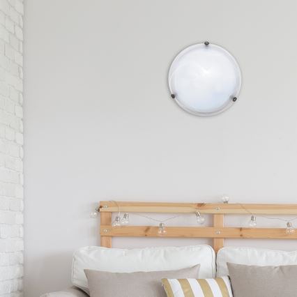 Okrúhle stropné svietidlo je pripevnené na stene nad posteľou s dreveným čelom a ozdobnými svetelnými bodkami. Svietidlo je biele a má kovový rám.