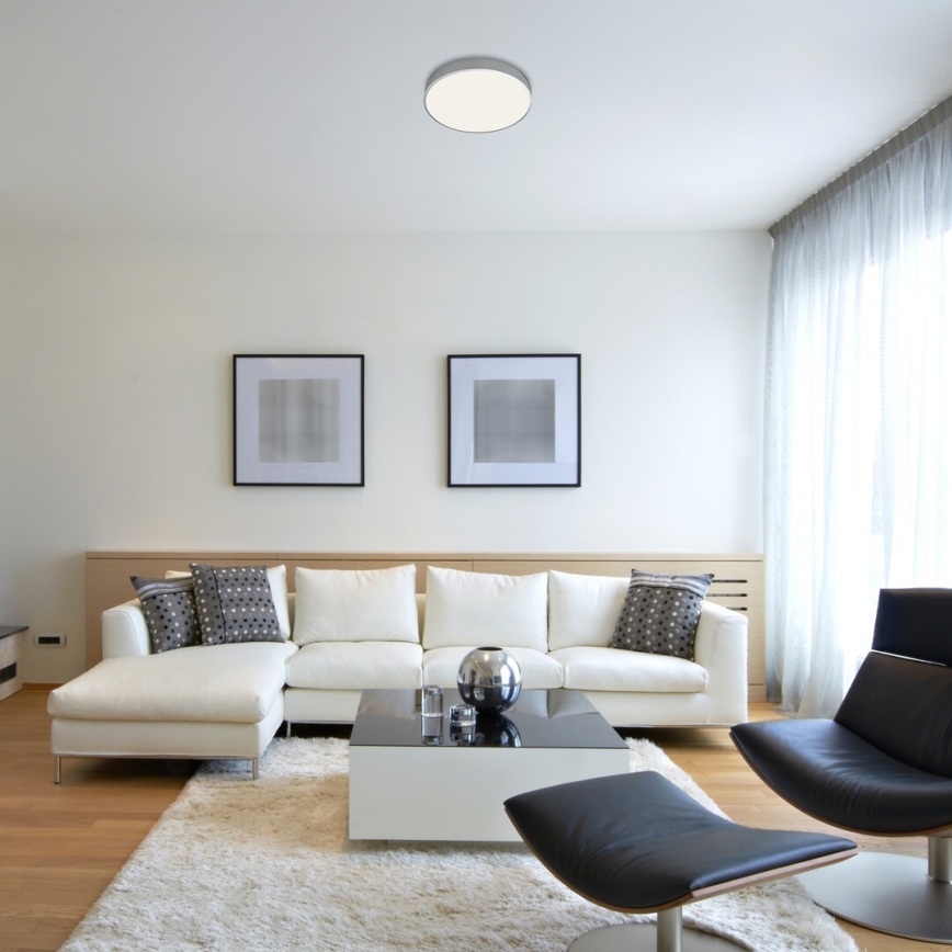 Svietidlo osvetľuje obývaciu izbu. Biela sedačka s rohovou časťou je umiestnená v miestnosti s bielymi stenami, svetlými závesmi a dvoma obrázkami.