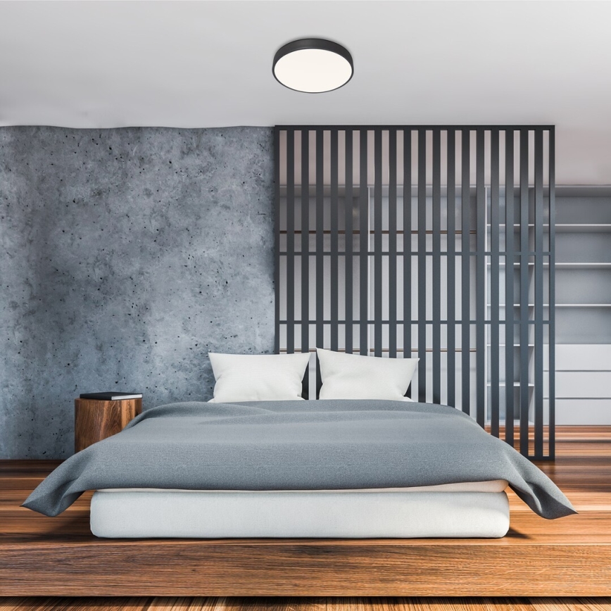 Svietidlo svieti v spálni. Drevená posteľ s šedou prikrývkou stojí pred priečkou. Stena je z betónu.