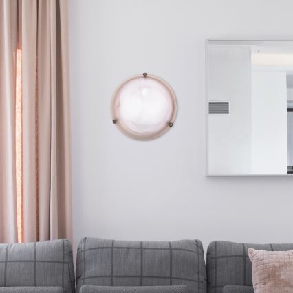 Okrúhla nástenná lampa svieti na stene obývacej izby. Lampa je umiestnená medzi šedou pohovkou a zrkadlom. Blízko je béžová záclona.