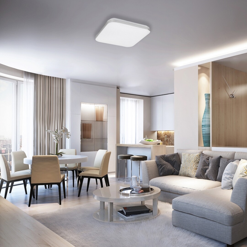 Štvorcové stropné svietidlo osvetľuje moderný obývací priestor. Svietidlo je umiestnené v strede miestnosti, ktorá obsahuje jedálenský stôl so stoličkami a rohovú sedačku.