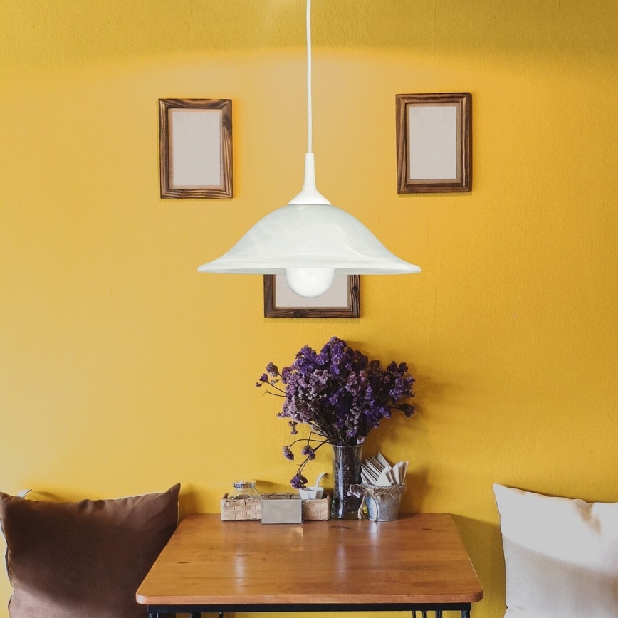 Biela závesná lampa svieti nad stolom. Na stole je váza s fialovými kvetmi, obrúsky a iné drobné predmety. Stôl stojí v miestnosti so žltými stenami a dvoma obrázkami v drevených rámoch.