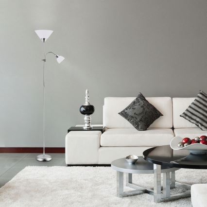 Stojacia lampa s dvoma tienidlami stojí v obývacej izbe s bielou sedačkou a dvoma konferenčnými stolíkmi. Lampa je strieborná, tienidlá sú biele. V pozadí je svetlošedá stena.
