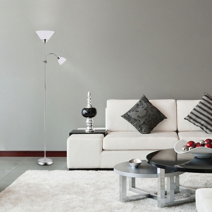 Stojacia lampa s dvoma tienidlami stojí v obývacej izbe s bielou sedačkou a dvoma konferenčnými stolíkmi. Lampa je strieborná, tienidlá sú biele. V pozadí je svetlošedá stena.