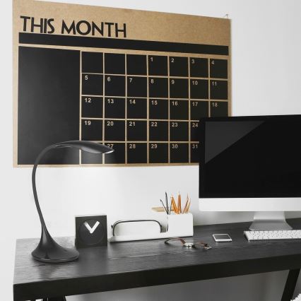 Stolná lampa osvetľuje pracovný stôl s počítačom, kalendárom na stene s nápisom „THIS MONTH“ a rôznymi kancelárskymi potrebami.
