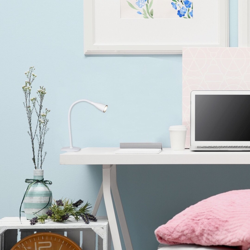 Biela stolová lampa svieti na bielom stole. Na stole je notebook a pohár. Pozadie tvorí svetlomodrá stena.