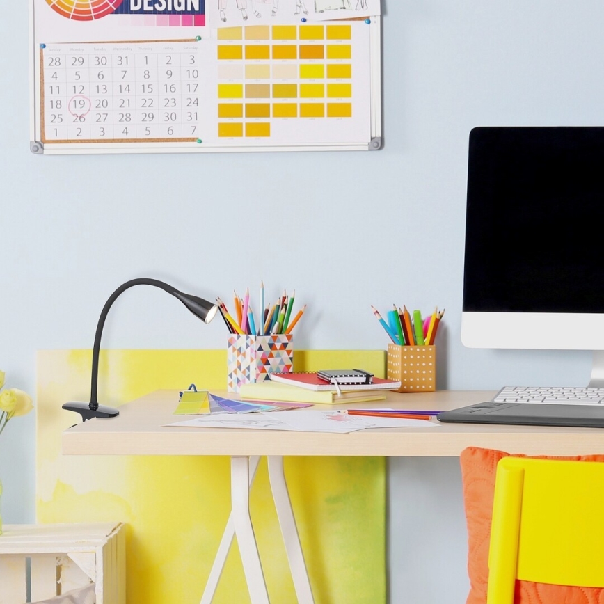 Stolová lampa s klipom osvetľuje písací stôl. Na stole sú ceruzky, zošity, počítač a grafický tablet. Stena za stolom je svetlomodrá a visí na nej kalendár.