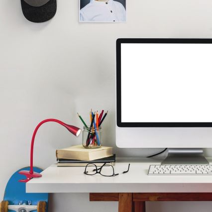 Červená stolná lampa so svorkou pripevnená na stôl svieti. Na stole je počítač, klávesnica, ceruzky v sklenenej nádobe, okuliare a knihy. Pozadie tvorí biela stena.