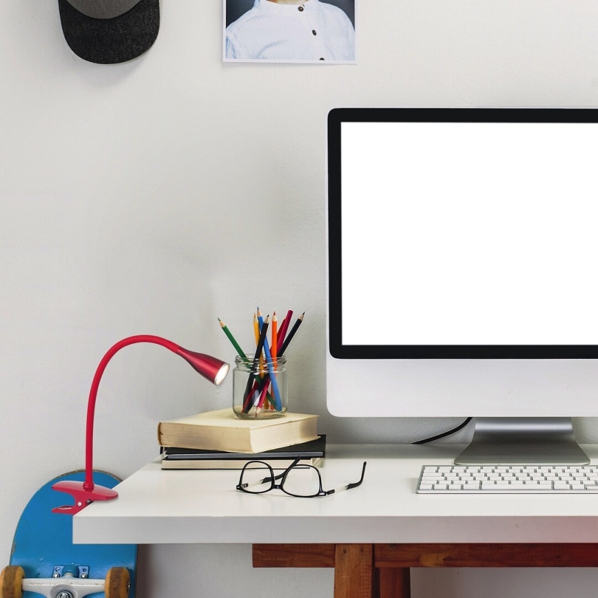 Červená stolná lampa so svorkou pripevnená na stôl svieti. Na stole je počítač, klávesnica, ceruzky v sklenenej nádobe, okuliare a knihy. Pozadie tvorí biela stena.