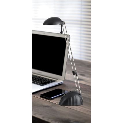 Stolová lampa s čiernou tienidlom a kovovými ramenami stojí na drevenom stole vedľa notebooku a mobilného telefónu.