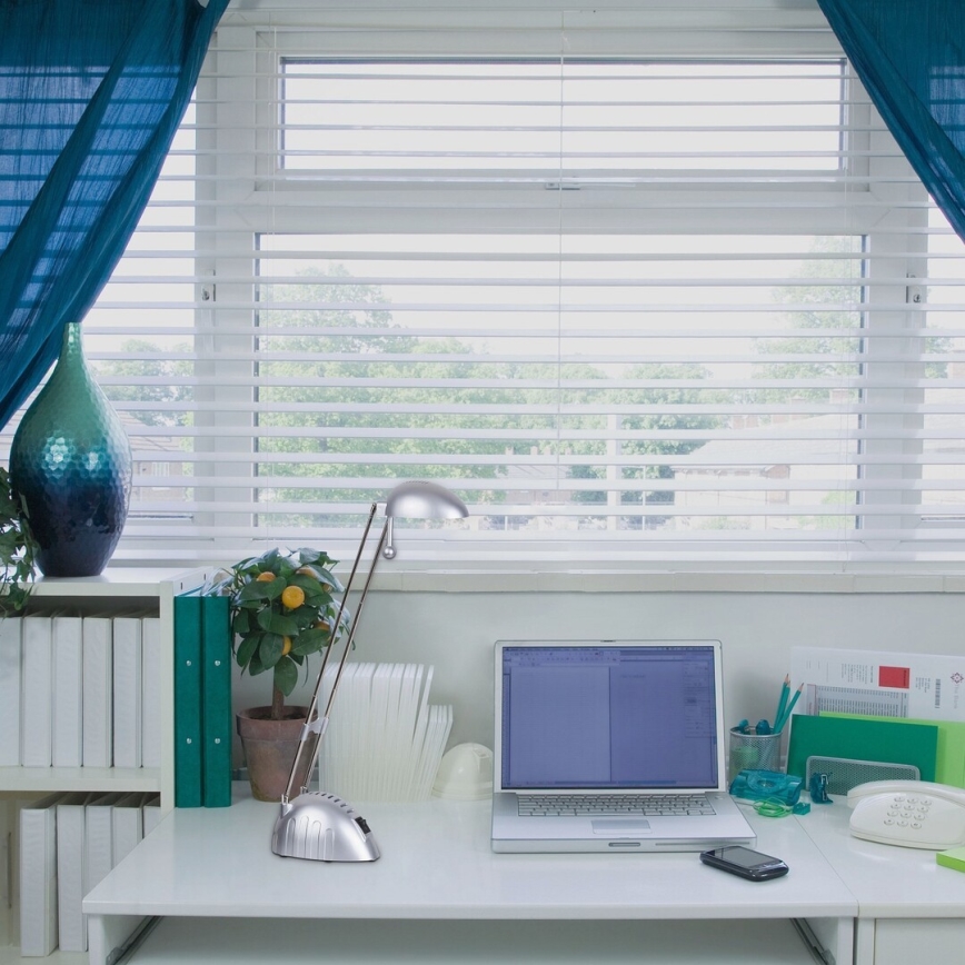 Stolová lampa svieti na notebook na bielom stole. Za stolom je okno s bielymi žalúziami a modrými závesmi. Na stole sú kancelárske potreby a kvetináč s citrusovým stromčekom.