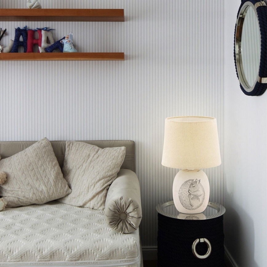 Stolová lampa s obrázkom veveričky stojí na nočnom stolíku. Lampa je umiestnená v detskej izbe popri pohovke s vankúšmi. Steny sú potiahnuté pruhovanou tapetou.