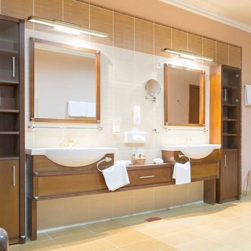 Dve umývadlá s drevenými skrinkami sa nachádzajú v kúpeľni s dlaždicami. Nad umývadlami sú zrkadlá a osvetlenie. V pozadí je drevená skriňa.