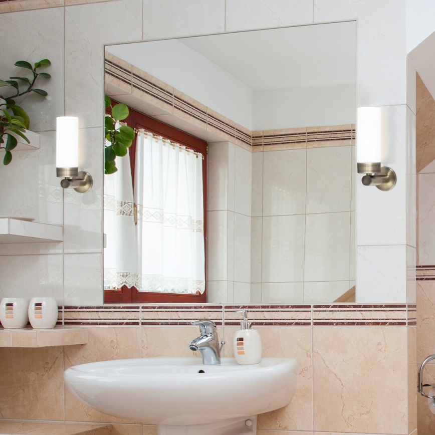 Dve nástenné svietidlá svietia v kúpeľni s umývadlom a zrkadlom. Svietidlá sú pripevnené na kachličkách. Zrkadlo odráža časť kúpeľne.