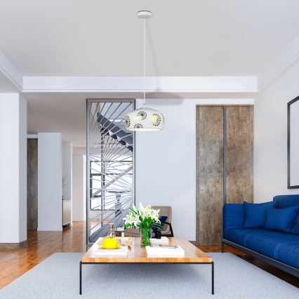 Závesné svietidlo s bielym tienidlom a vzorom kvetov visí v modernom interiéri. Svietidlo osvetľuje obývaciu izbu s drevenou podlahou a nízkym konferenčným stolíkom. V pozadí je vidieť schodisko za kovovou priečkou.