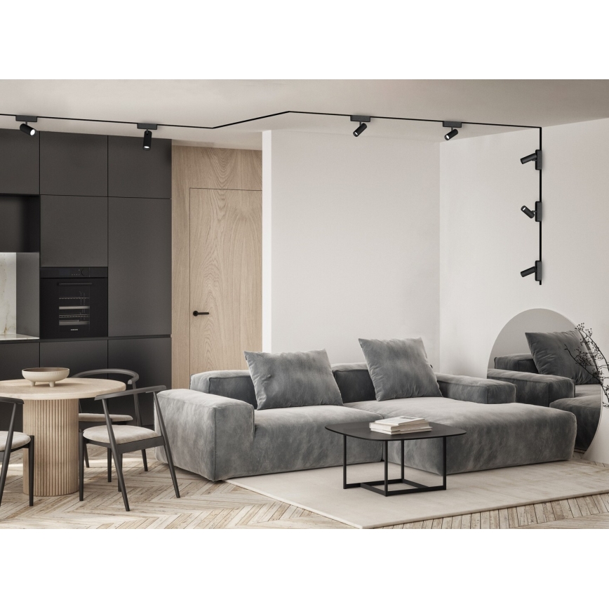 Šedá rohová sedačka stojí v obývacej izbe s drevenou podlahou. Nad ňou je systém bodových svietidiel na koľajniciach. V pozadí je kuchyňa s tmavými skrinkami a jedálenským stolom.