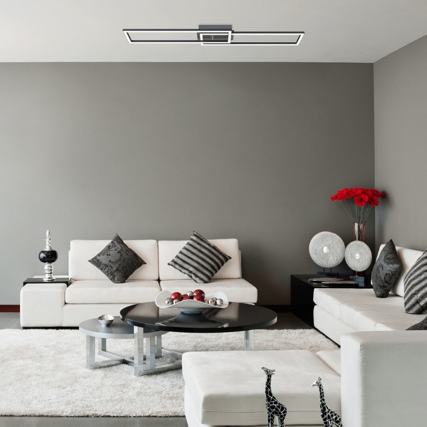 Stropné svietidlo svieti v modernej obývačke. Svietidlo je dlhé, obdĺžnikové, s dvoma vnorenými svetlami. Obývačka obsahuje bielu sedačku a konferenčný stolík.