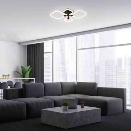 Svietidlo svieti v modernom interiéri. Štvorramenné stropné svietidlo s hranatými tienidlami je umiestnené nad rozsiahlou tmavosivou sedačkou a nízkym kamenným stolíkom v miestnosti s veľkými oknami s výhľadom na mesto.