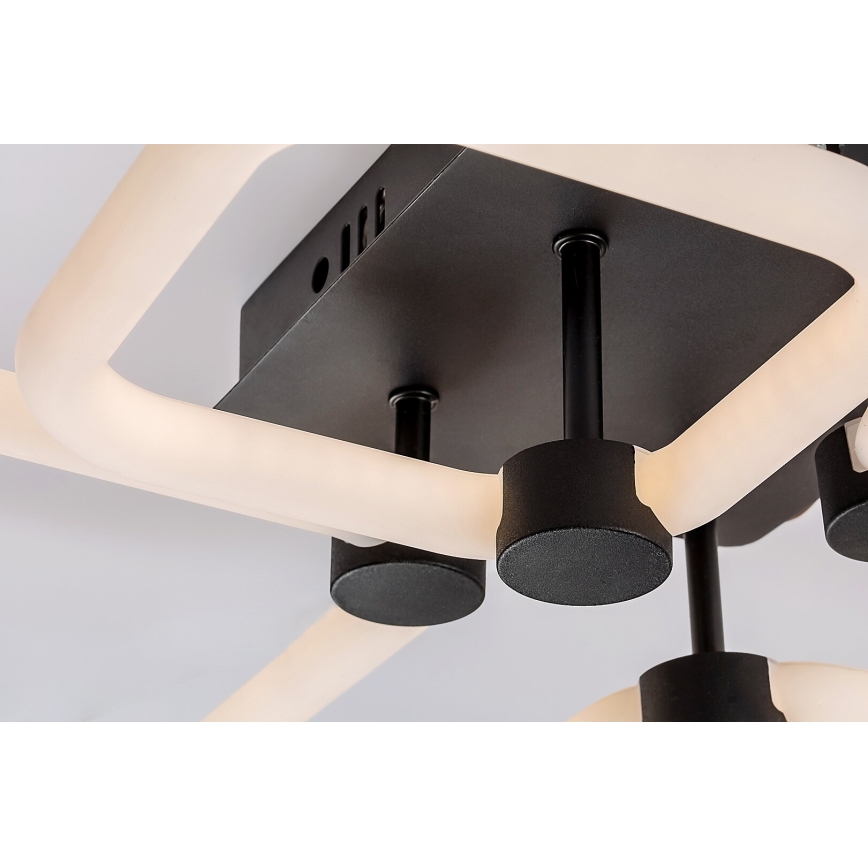 Svietidlo svieti. Má čierny štvorcový základ a biele LED prvky v tvare prepojených oblúkov. Je pripevnené k stropu.