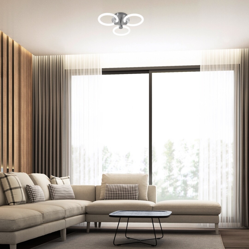 Trojramenné stropné svietidlo s kruhovými prvkami je osvetlené v modernej obývačke s rohovou sedačkou, konferenčným stolíkom a veľkým oknom s priesvitnými závesmi. Stenu čiastočne tvorí drevený obklad.