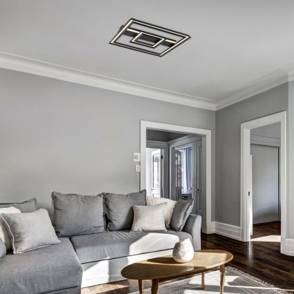 Svetlo štvorcového tvaru s vnútornými geometrickými prvkami je pripevnené na strope. Svieti v obývacej izbe s rohovou sedačkou a konferenčným stolíkom. Steny a podlaha sú sivé a hnedé.