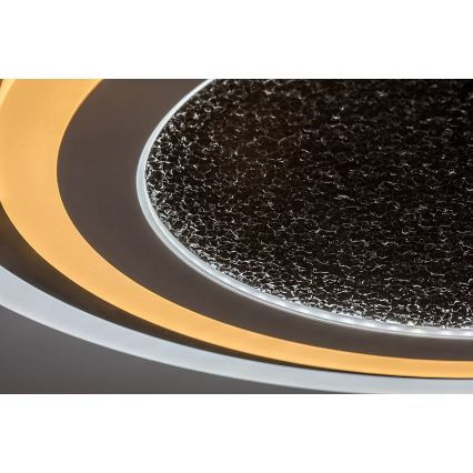 Svietidlo svieti. Má okrúhly tvar s vnútorným tmavosivým sklom s drsným povrchom a dvojitým vonkajším okrajom s teplým bielym LED osvetlením. Je namontované na tmavom pozadí.