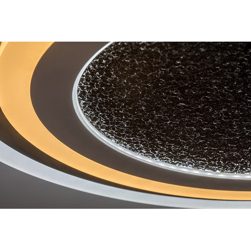 Svietidlo svieti. Má okrúhly tvar s vnútorným tmavosivým sklom s drsným povrchom a dvojitým vonkajším okrajom s teplým bielym LED osvetlením. Je namontované na tmavom pozadí.