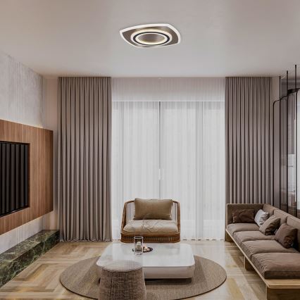 Stropné svietidlo s dvojitým kruhovým dizajnom svieti v moderne zariadenej obývacej izbe. Izba obsahuje sedačku, kreslo, konferenčný stolík a dlhé závesy. Podlaha je drevená.