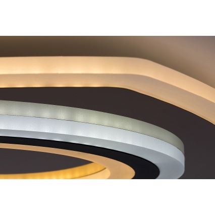 Svietidlo svieti v interiéri. Má okrúhly tvar s viacerými koncentrickými kruhmi v odtieňoch sivej, bielej a zlatej. Vonkajší okraj je osvetlený.