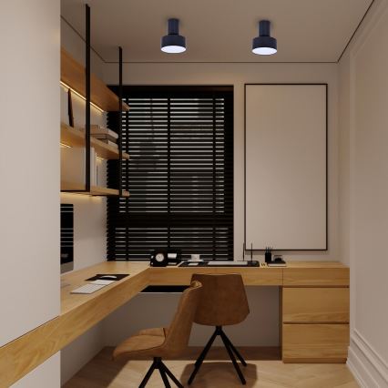 Drevený písací stôl s dvoma hnedými stoličkami je umiestnený v kancelárii. Nad stolom visí prázdny obraz. Na stene sú drevené police a čierne žalúzie na okne. Na strope sú dve tmavomodré svietidlá.