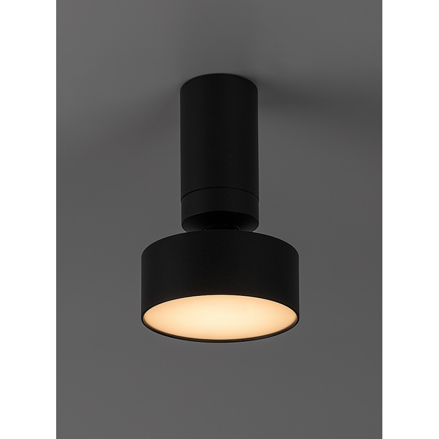 Čierna stropná lampa s valcovým telom a kruhovým difúzorom svieti. Difúzor je mierne otočený od stropu. Lampa je pripevnená k stropu jednotného svetlohnedého odtieňa.