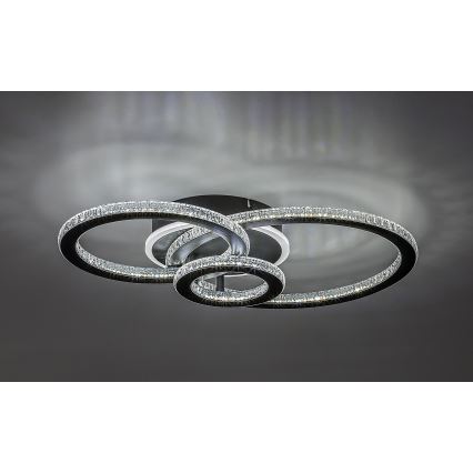 Svietidlo s tromi prepletenými kruhmi osadenými kryštálmi svieti na strope. Kruhy sú tmavosivé, kryštály priehľadné. Svetlo je umiestnené v tmavosivej miestnosti.