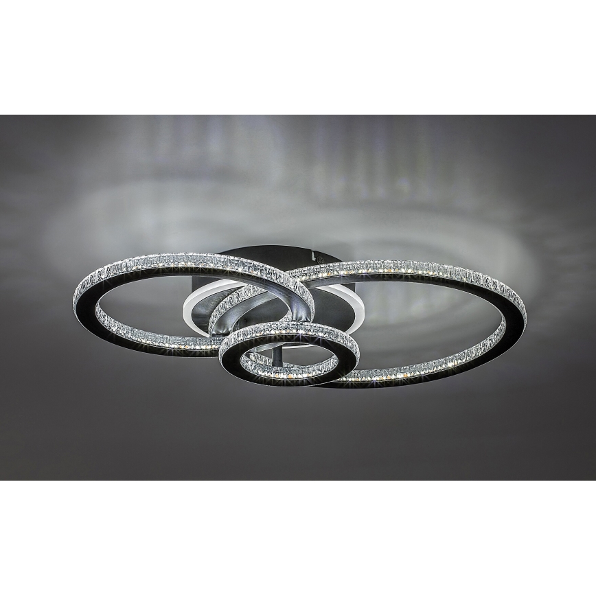 Svietidlo s tromi prepletenými kruhmi osadenými kryštálmi svieti na strope. Kruhy sú tmavosivé, kryštály priehľadné. Svetlo je umiestnené v tmavosivej miestnosti.