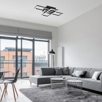 Moderná stropná lampa s geometrickým dizajnom svieti v obývacej izbe. Šedá rohová sedačka s vankúšmi stojí na svetlom koberci pred oknom s balkónom. V pozadí je jedálenský stôl a stoličky.