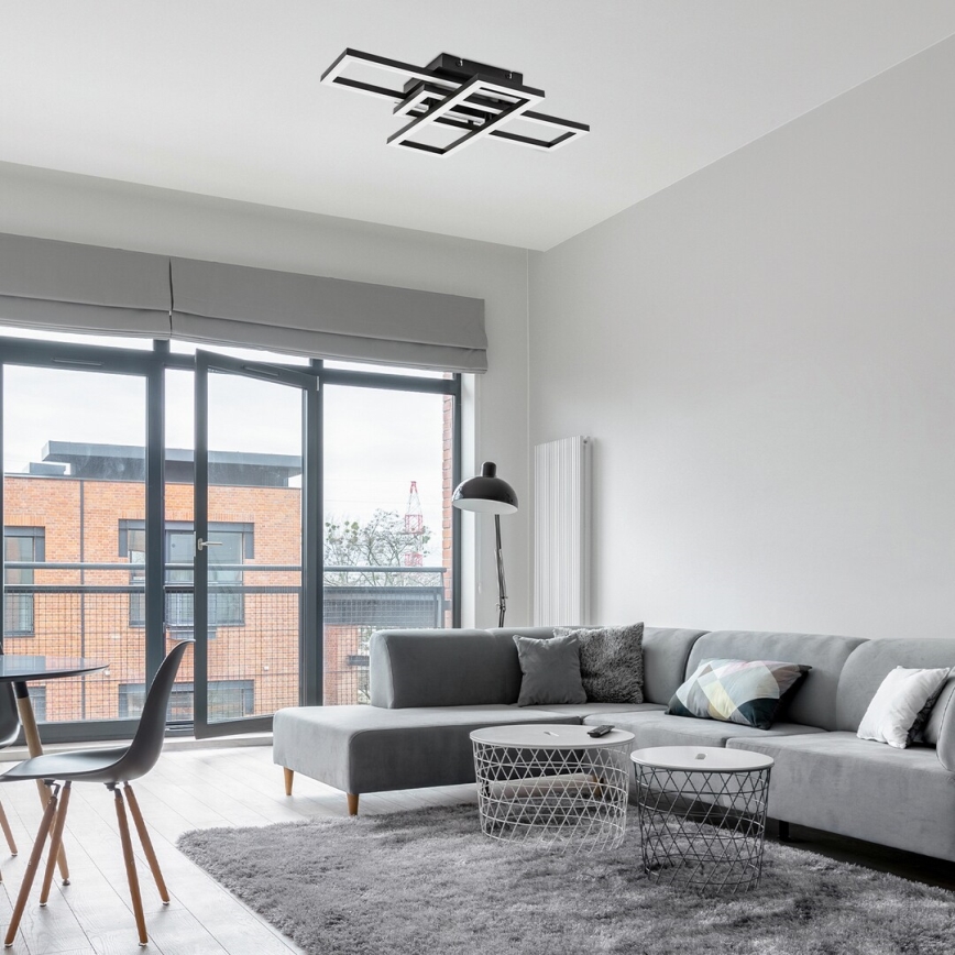 Moderná stropná lampa s geometrickým dizajnom svieti v obývacej izbe. Šedá rohová sedačka s vankúšmi stojí na svetlom koberci pred oknom s balkónom. V pozadí je jedálenský stôl a stoličky.