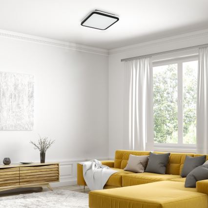 Štvorcový stropný svietidlo s bielym difúzorom a čiernym rámom je osvetlené v svetlej obývačke s žltou sedačkou a veľkým oknom.