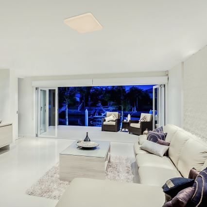 Štvorcové stropné svietidlo svieti v obývacej izbe. Svetlo osvetľuje svetlý nábytok, koberec a výhľad cez posuvné dvere na nočnú vodnú plochu s člnmi.