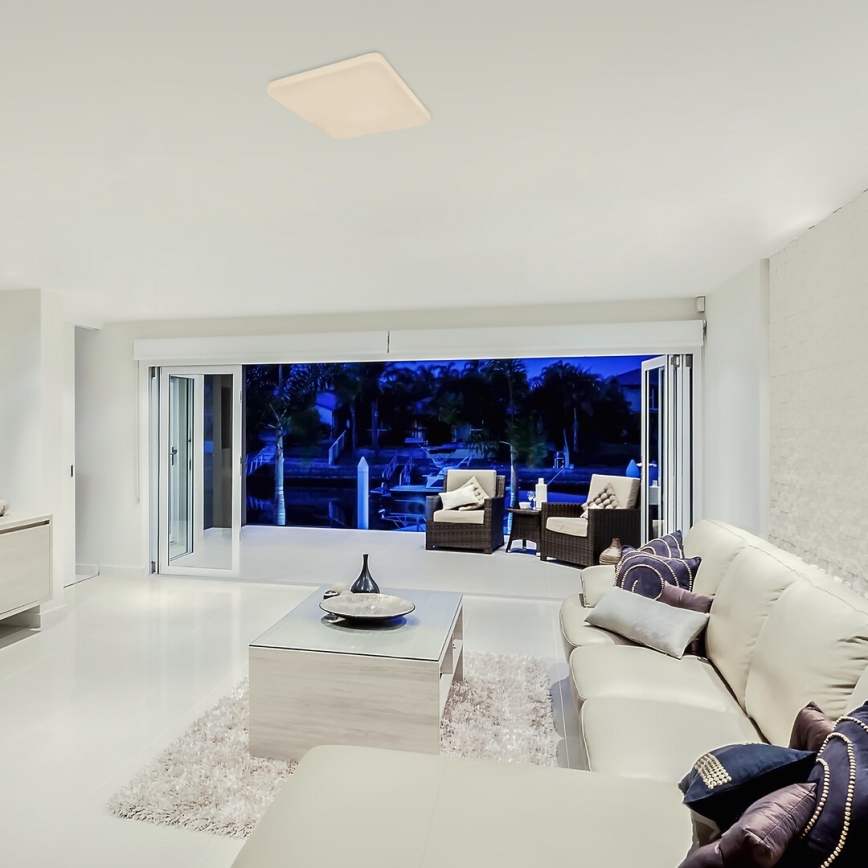 Štvorcové stropné svietidlo svieti v obývacej izbe. Svetlo osvetľuje svetlý nábytok, koberec a výhľad cez posuvné dvere na nočnú vodnú plochu s člnmi.