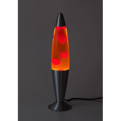 Lávová lampa svieti. V oranžovej tekutine plávajú červené kvapky. Stojí na kovovom podstavci na tmavosivej ploche.