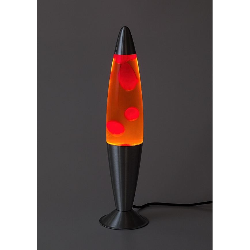 Lávová lampa svieti. V oranžovej tekutine plávajú červené kvapky. Stojí na kovovom podstavci na tmavosivej ploche.