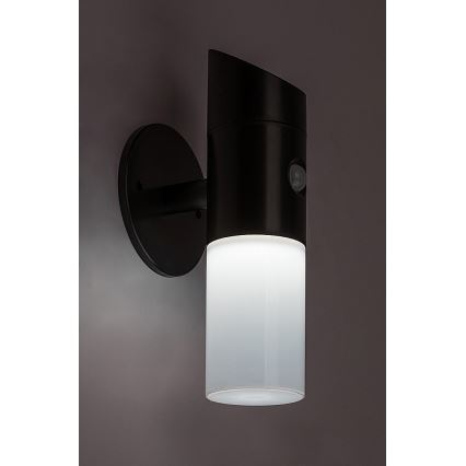 Čierna nástenná lampa s pohybovým senzorom svieti. Svetlo vyžaruje z valcovitého mliečneho difúzora. Lampa je namontovaná na stene.