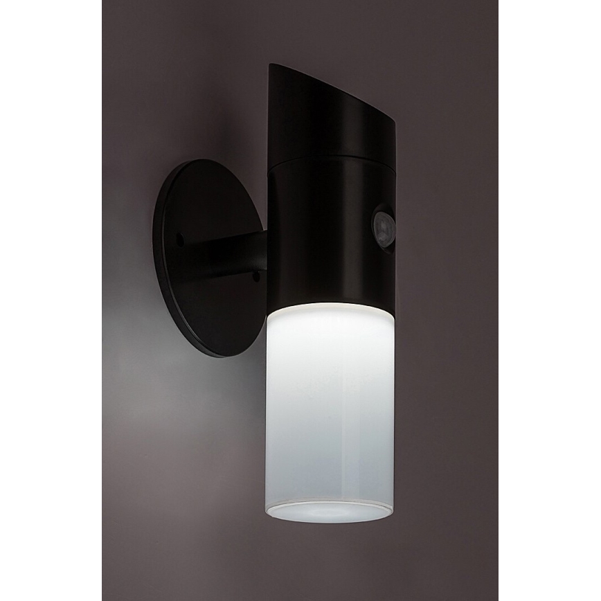 Čierna nástenná lampa s pohybovým senzorom svieti. Svetlo vyžaruje z valcovitého mliečneho difúzora. Lampa je namontovaná na stene.