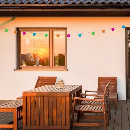 Drevený záhradný nábytok stojí na terase. Farebné papierové lampióny visia na šnúre pred oknom s odrazom západu slnka.