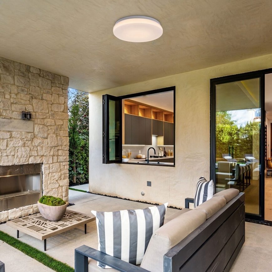 Svetlo svieti na vonkajšej terase s kamenným krbom, sedacou súpravou a výhľadom na kuchyňu cez otvorené okno.