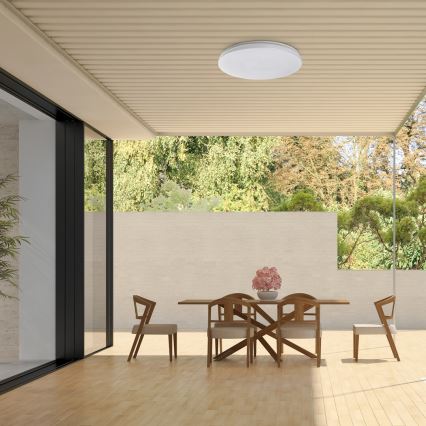 Svetlo svieti na drevenom stole a stoličkách v jedálni pod vonkajším prístreškom s výhľadom na zeleň.