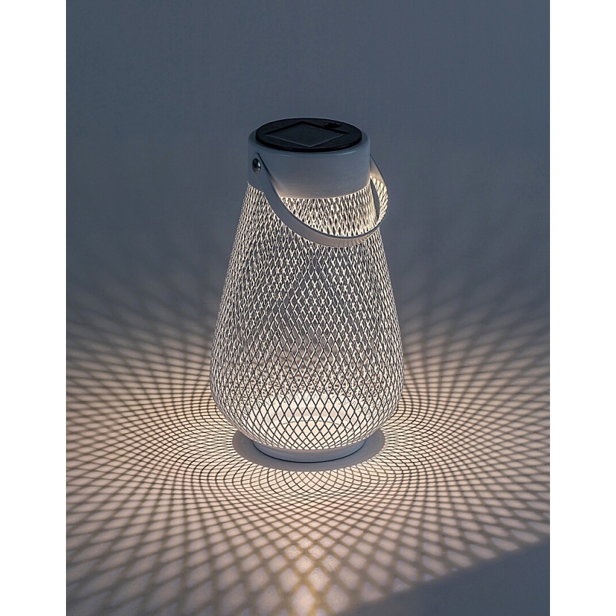 Solárna lampa svieti. Biela kovová konštrukcia s rúčkou a sieťovaným telom vytvára na podložke svetelný vzor.