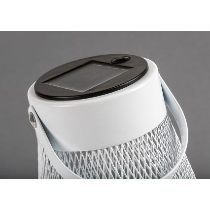 Biela solárna lampa s rúčkou a sieťovaným telom. Solárny panel je na vrchu. Lampa je umiestnená na sivom pozadí.