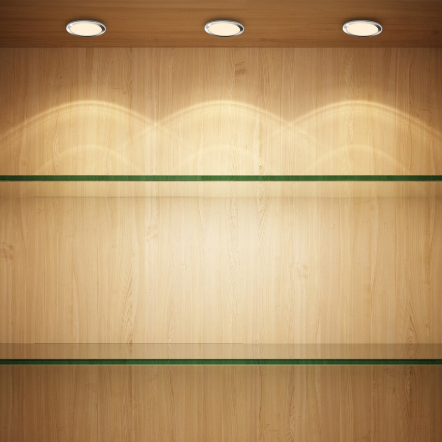 Dve prázdne sklenené police sú umiestnené v drevenej vitríne osvetlenej tromi stropnými svetlami. Svetlá vytvárajú na drevenom povrchu osvetlené oblasti.