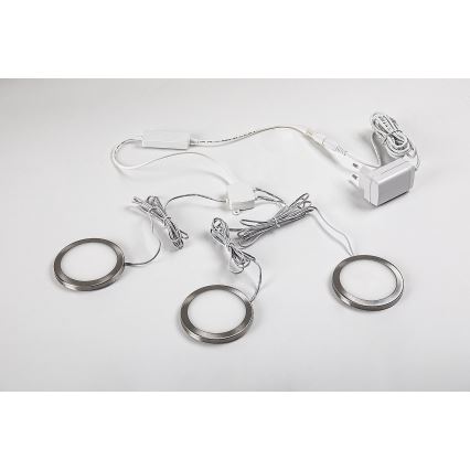 Tri okrúhle LED svetlá s kovovými rámami sú umiestnené na svetlom povrchu. Sú prepojené káblami a napájacím zdrojom s európskou zástrčkou.