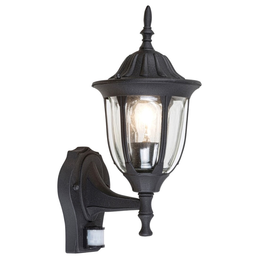 Čierna nástenná lampa s pohybovým senzorom svieti. Jej sklenená časť je oválna, kovová časť je zdobená. Lampa je pripevnená na stenu.